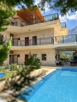 Un edificio de dos plantas con piscina, zona de estar al aire libre y montañas al fondo.