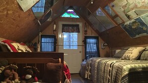 Een gezellig zolderkamer met een schuine daklijn, een houten deur, twee ramen, een bed met sprei, een stoel met knuffeldieren en een klein vloerkleed.