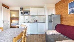 Eine kleine Küche mit weißen Schränken, einer Mikrowelle, einem Kühlschrank und einem Geschirrspüler. Ein Holztisch mit Stühlen, ein Sofa mit roter Decke und eine Holzvertäfelung mit einem gerahmten Bild.