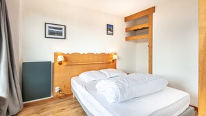 Ein ordentlich bezogenes Bett mit zwei Kissen und einer zusammengelegten Decke, ein hölzernes Kopfteil mit Wandlampen und ein kleines Regal darüber.