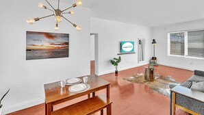 A modern living room with a wooden dining table, a sofa, a hanging light fixture, and a large window with blinds. - Garden Suite 1-hosted by Sweetstay (Miami Beach)