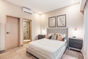 A bedroom with a large bed, a bedside table, a nightstand, an air conditioning unit, and a framed artwork on the wall.