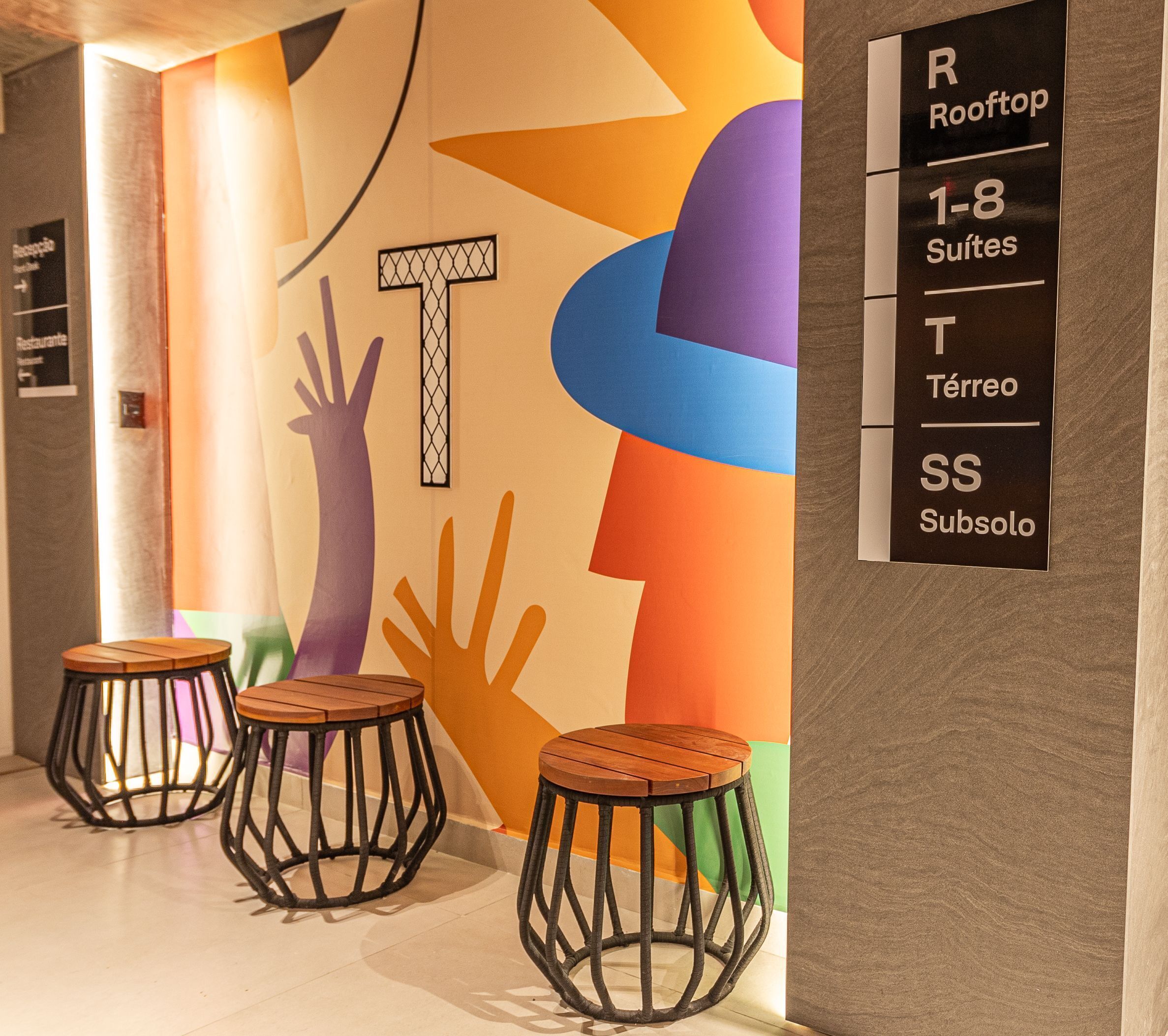 A hotel interior with a colorful wall design, three wooden stools, and a sign indicating room types.