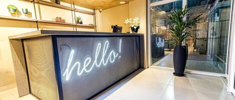 A reception desk with a 'hello!' sign, potted plants, and a view of the outside through large windows.