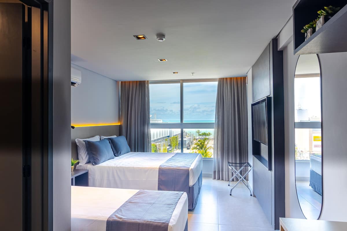 Ein Hotelzimmer mit zwei Betten, einem Fernseher und einem Balkon mit Blick auf Stadt und Meer.