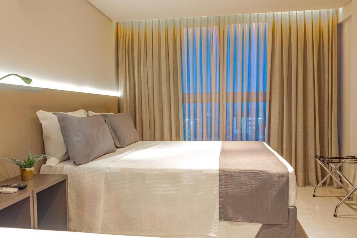 Ein Hotelzimmer mit einem Bett, einem Nachttisch mit Pflanze, einem kleinen Tisch mit Lampe und einem Fenster mit Vorhängen.