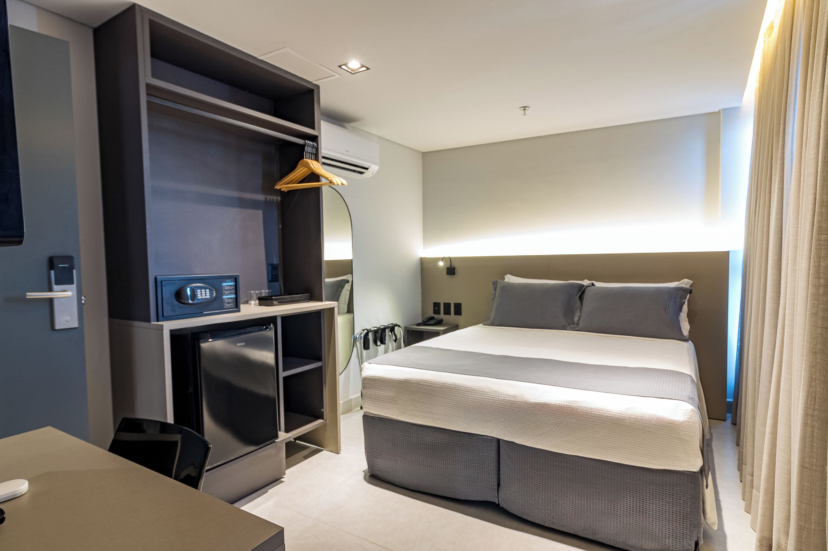 Una habitación de hotel moderna con una cama grande, un escritorio empotrado con microondas, un refrigerador pequeño y un armario con perchero.