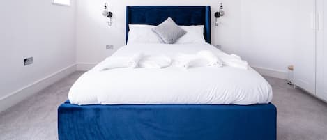 A neatly made bed with white linens and a blue headboard in a room with white walls and a carpeted floor.
