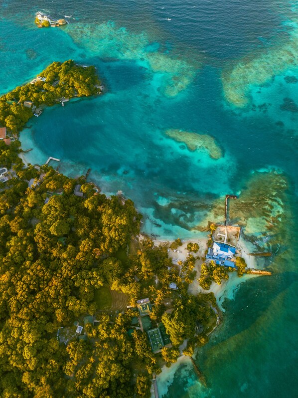 Luchtbeeld van een tropisch eiland met helder turquoise water, weelderige groene begroeiing en enkele gebouwen, waaronder een groot huis met een blauw dak.