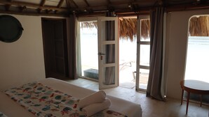 Una habitación con una cama, una mesita y una puerta que da a un espacio exterior con techo de paja.