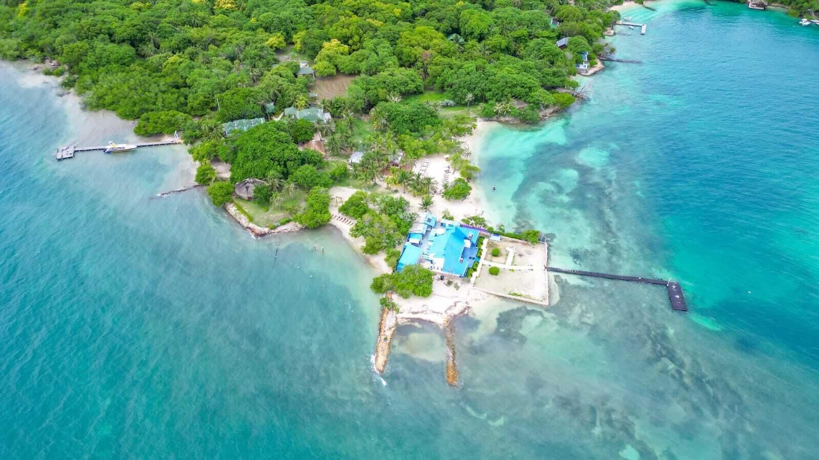An aerial view of a tropical island with a clear blue sea, a dock, and a building with a swimming pool.