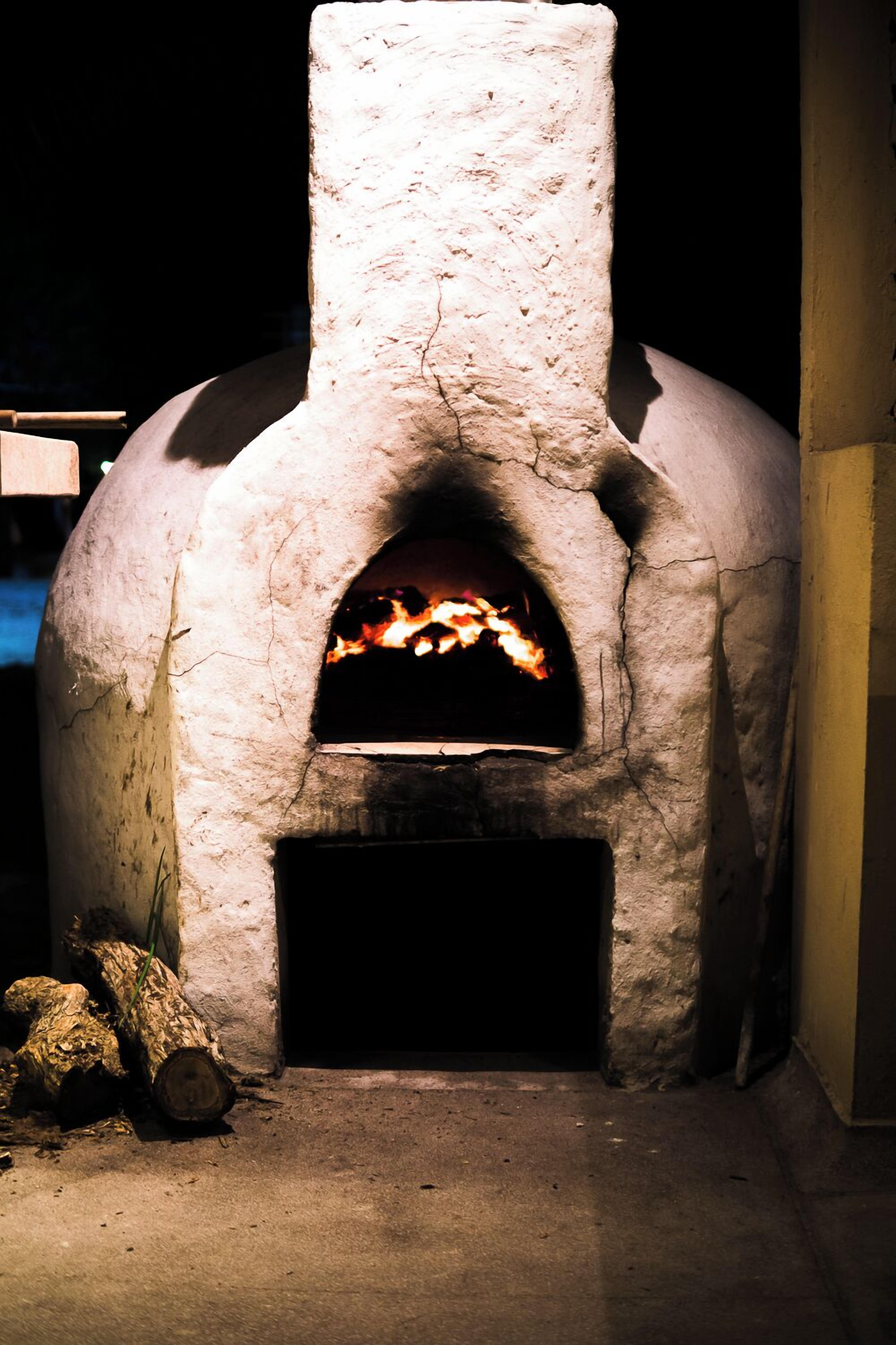 A stone-built outdoor pizza oven with a fire burning inside, logs placed beside it, and a dark background.