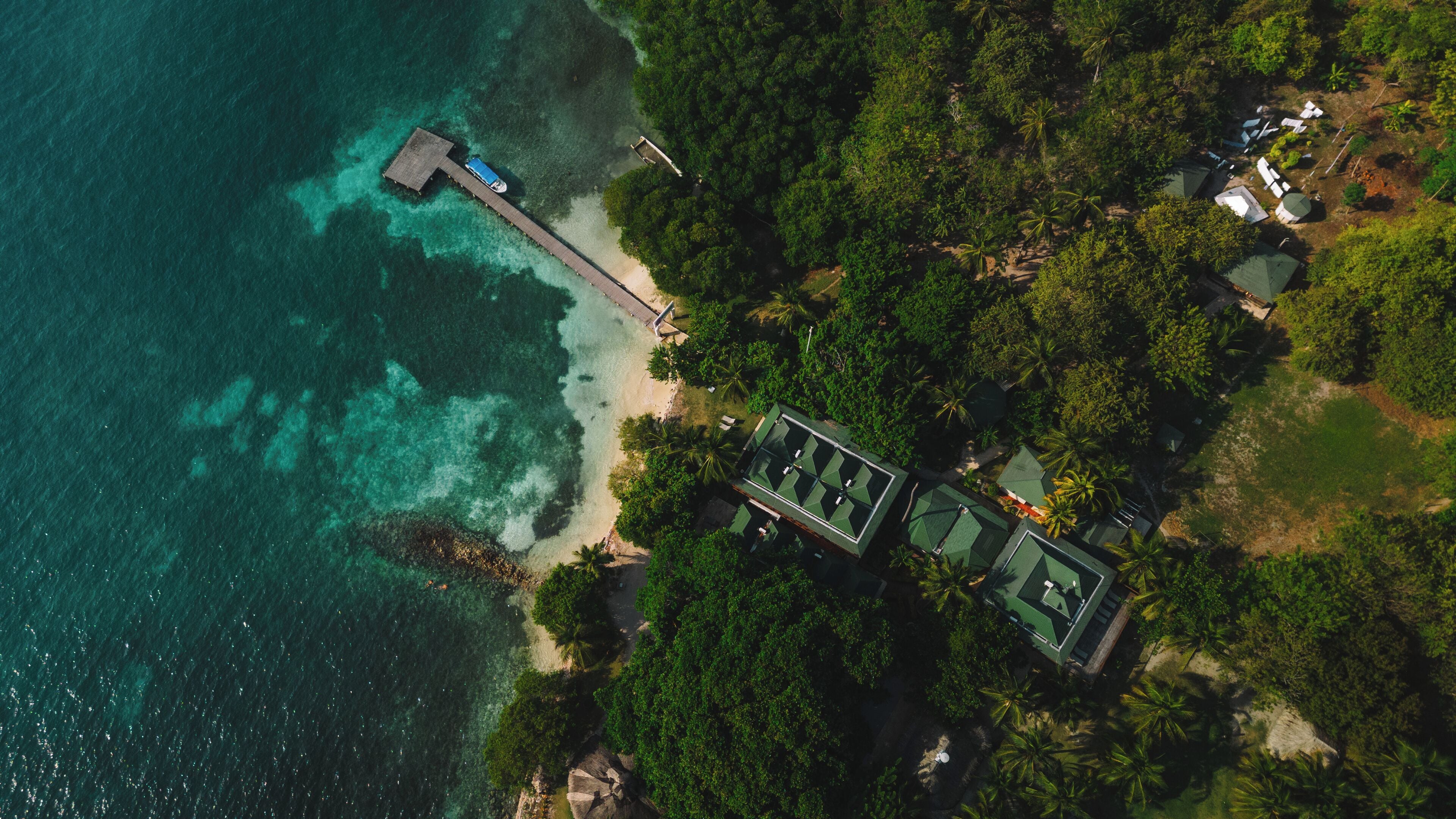 Vista aérea de un resort con un muelle que se adentra en aguas turquesas, rodeado de exuberante vegetación y un conjunto de edificios con tejados verdes.