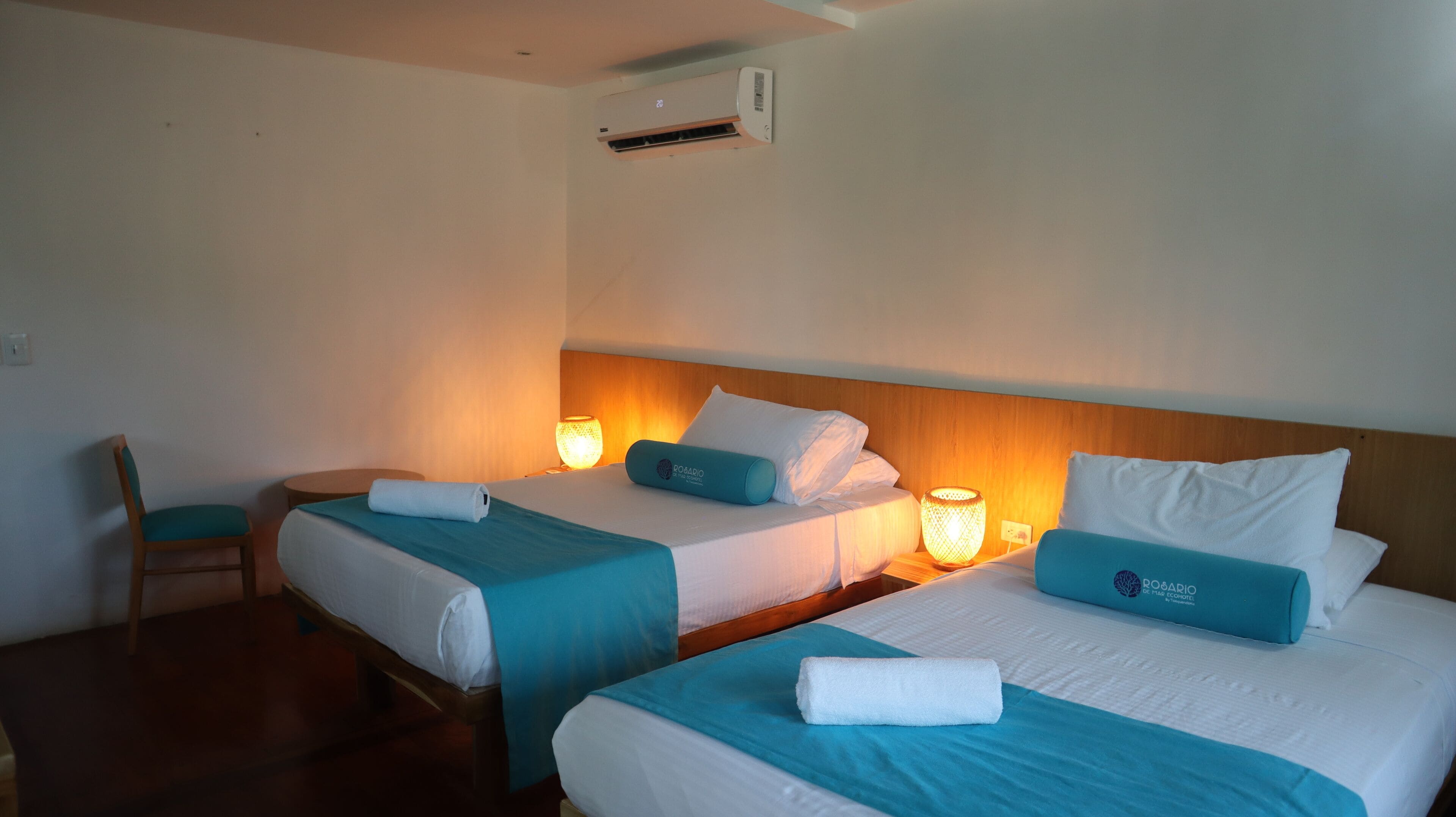 A hotel room with two beds, each with a blue bedspread and white pillows, a wooden headboard, and a wall-mounted air conditioner.