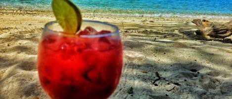 Un vaso de bebida roja con lima sobre una playa arenosa, con un océano azul cristalino y un barco a lo lejos.
