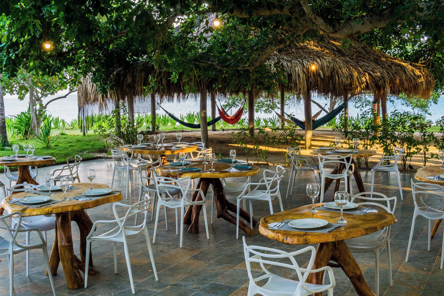 An outdoor dining area with round tables and white chairs, surrounded by trees and a thatched roof structure.