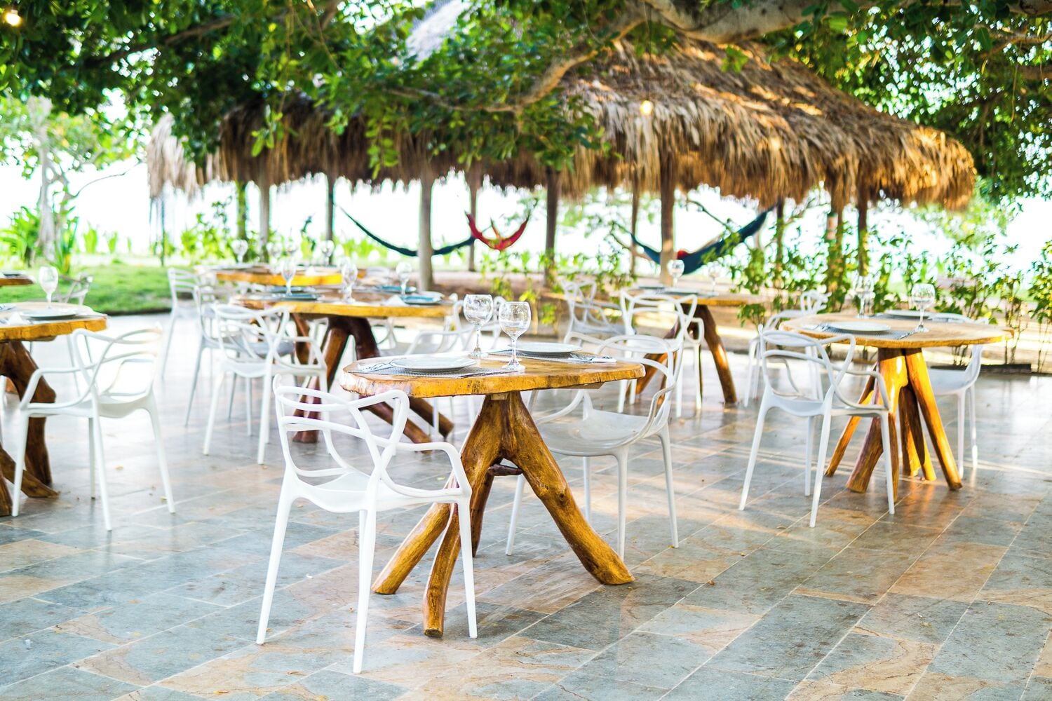 An outdoor dining area with round tables and white chairs, surrounded by greenery and a thatched roof structure.