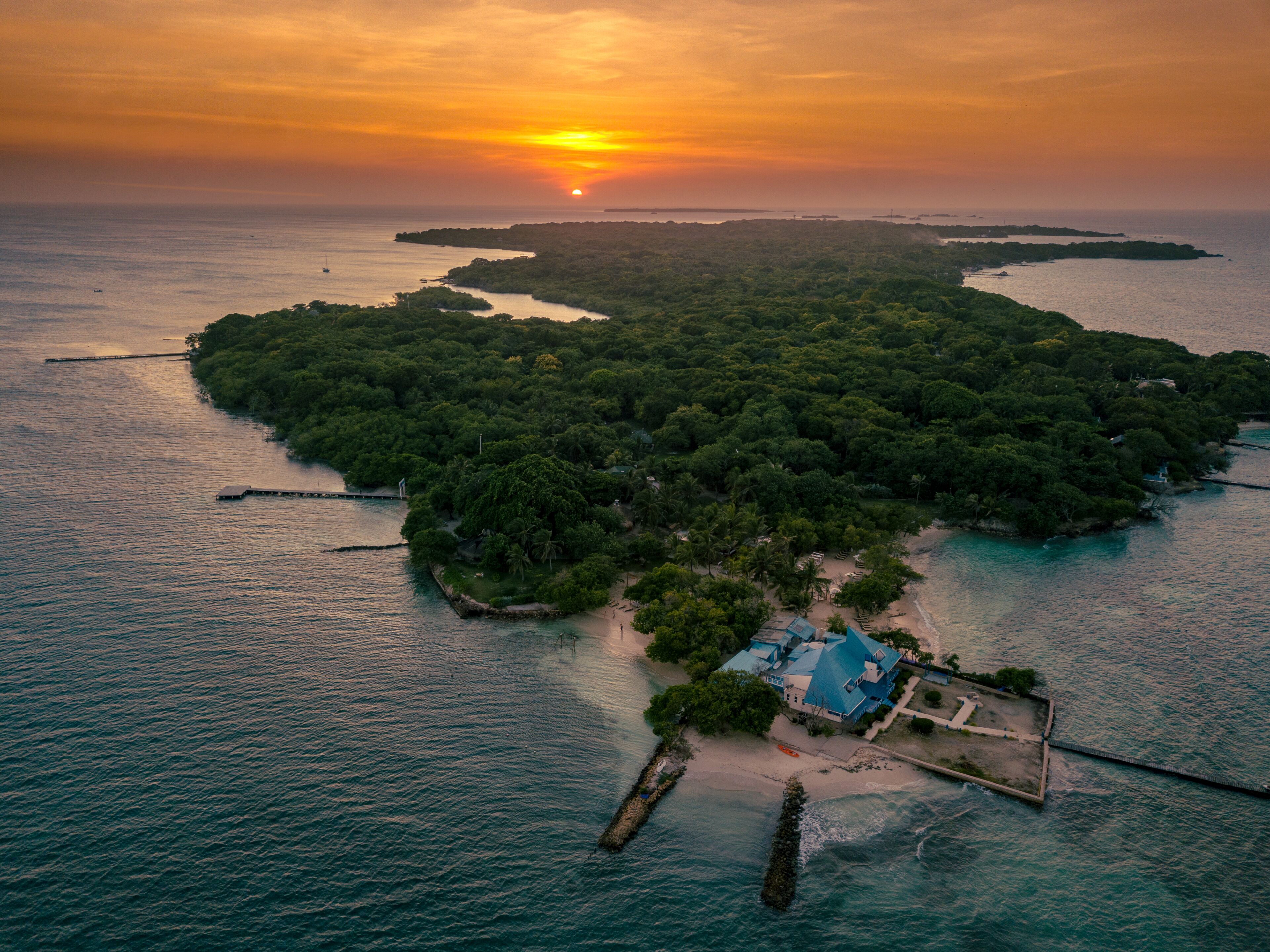 Vista aérea de una isla tropical con abundante vegetación, un pequeño muelle y un edificio con techo azul.