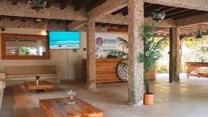 Zona de recepción con un mostrador de madera, un espejo grande y un televisor que muestra una escena de playa.