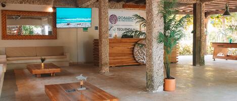 Zona de recepción con un mostrador de madera, un espejo grande y un televisor que muestra una escena de playa.