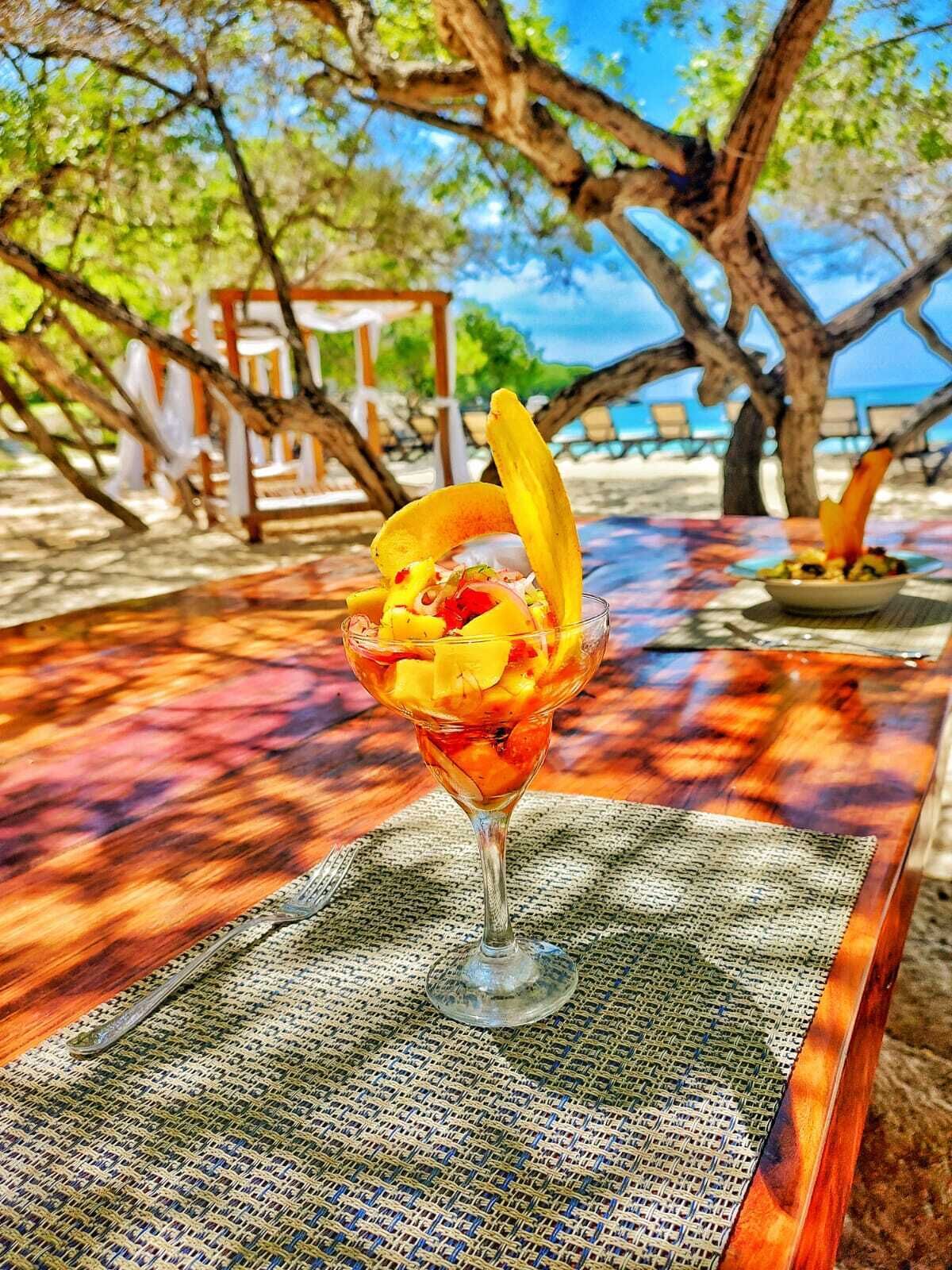 A tropical drink in a glass on a wooden table with a woven placemat, set against a beach backdrop with trees and a clear sky.