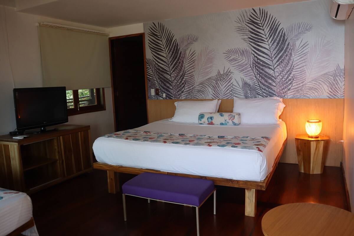 Habitación de hotel con cama, televisión, banco, mesita de noche con lámpara y pared con diseño de hojas.