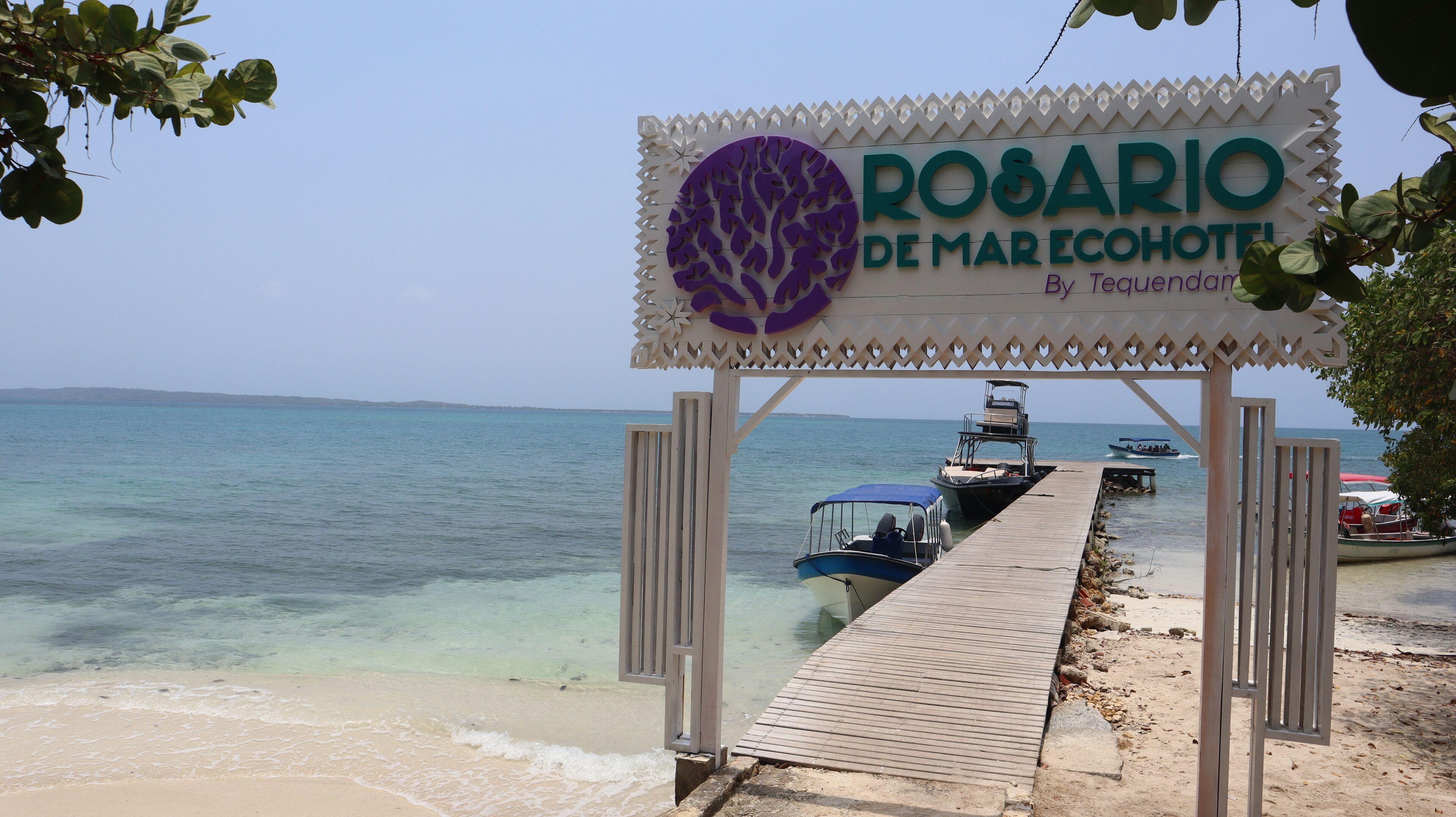 En träbrygga som leder till en strand, och där finns en skylt med texten ”Rosario de Mar Ecohotel”.