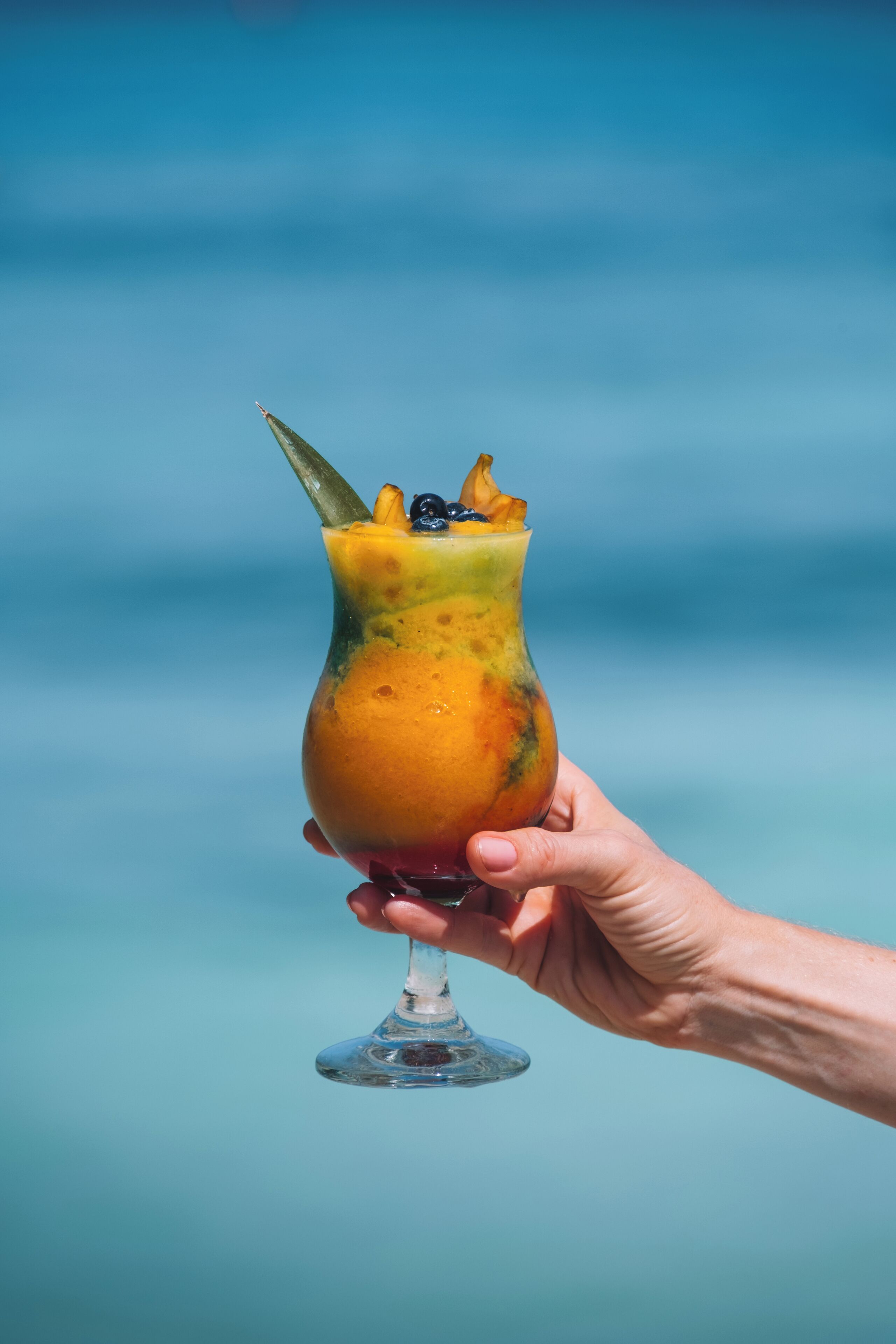 A hand holding a colorful drink with a tropical fruit garnish against a blue background.