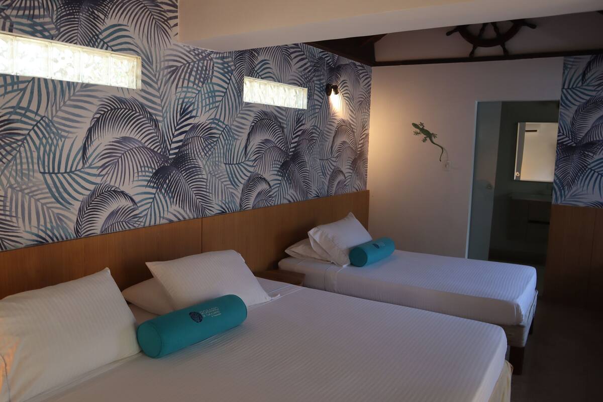 Habitación de hotel con dos camas, cada una con sábanas blancas y almohadas turquesa. La habitación presenta un papel tapiz con motivos de hojas tropicales y un cabecero de madera.