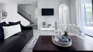 A modern living room with a dark sofa, a coffee table with decorative items, a staircase, a flat-screen TV, and a large window with curtains. - Balmoral Resort-213al 3 Bedroom Home by RedAwning (Haines City)