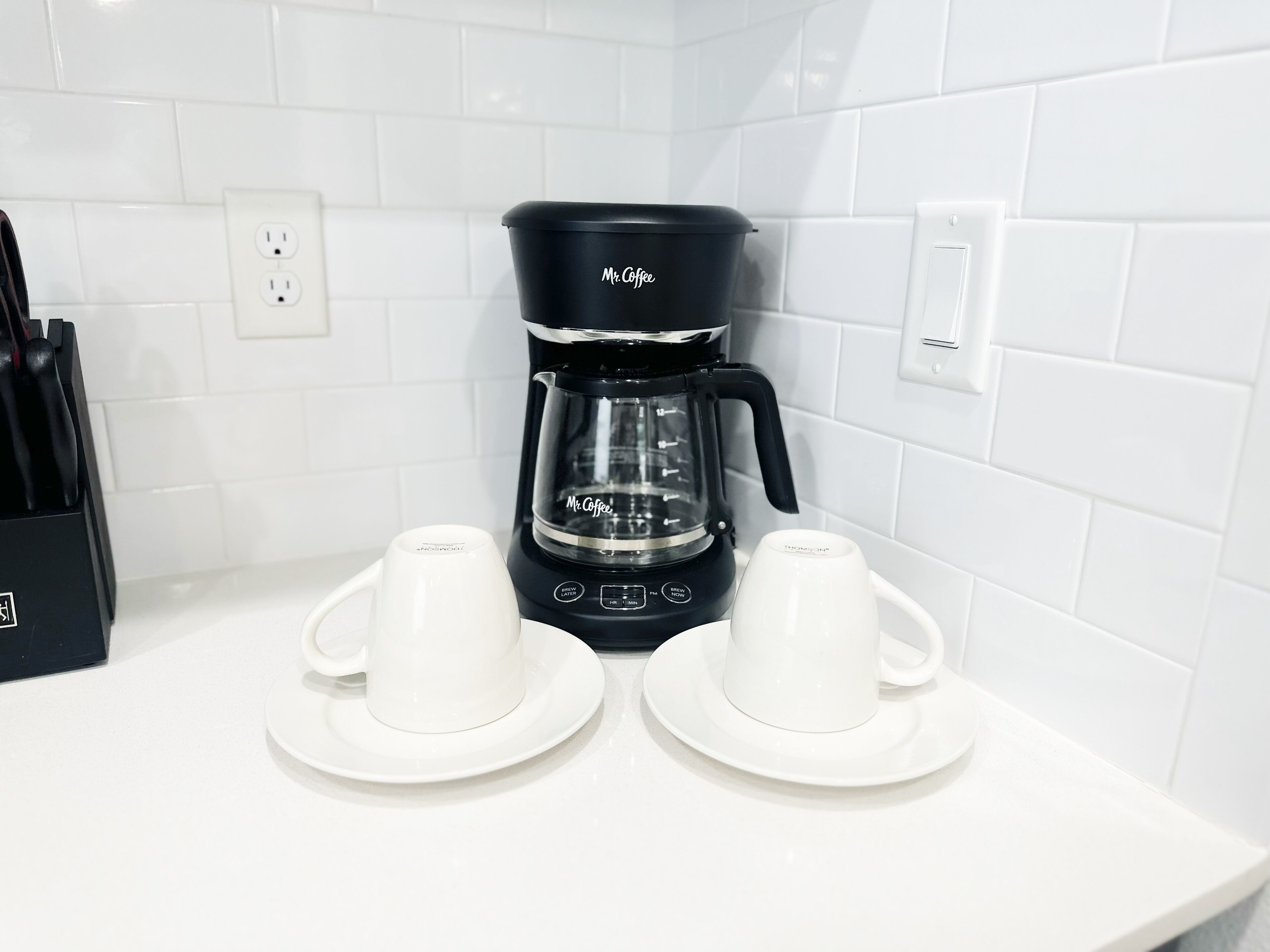 A black Mr. Coffee coffee maker on a white countertop with two white cups and saucers.