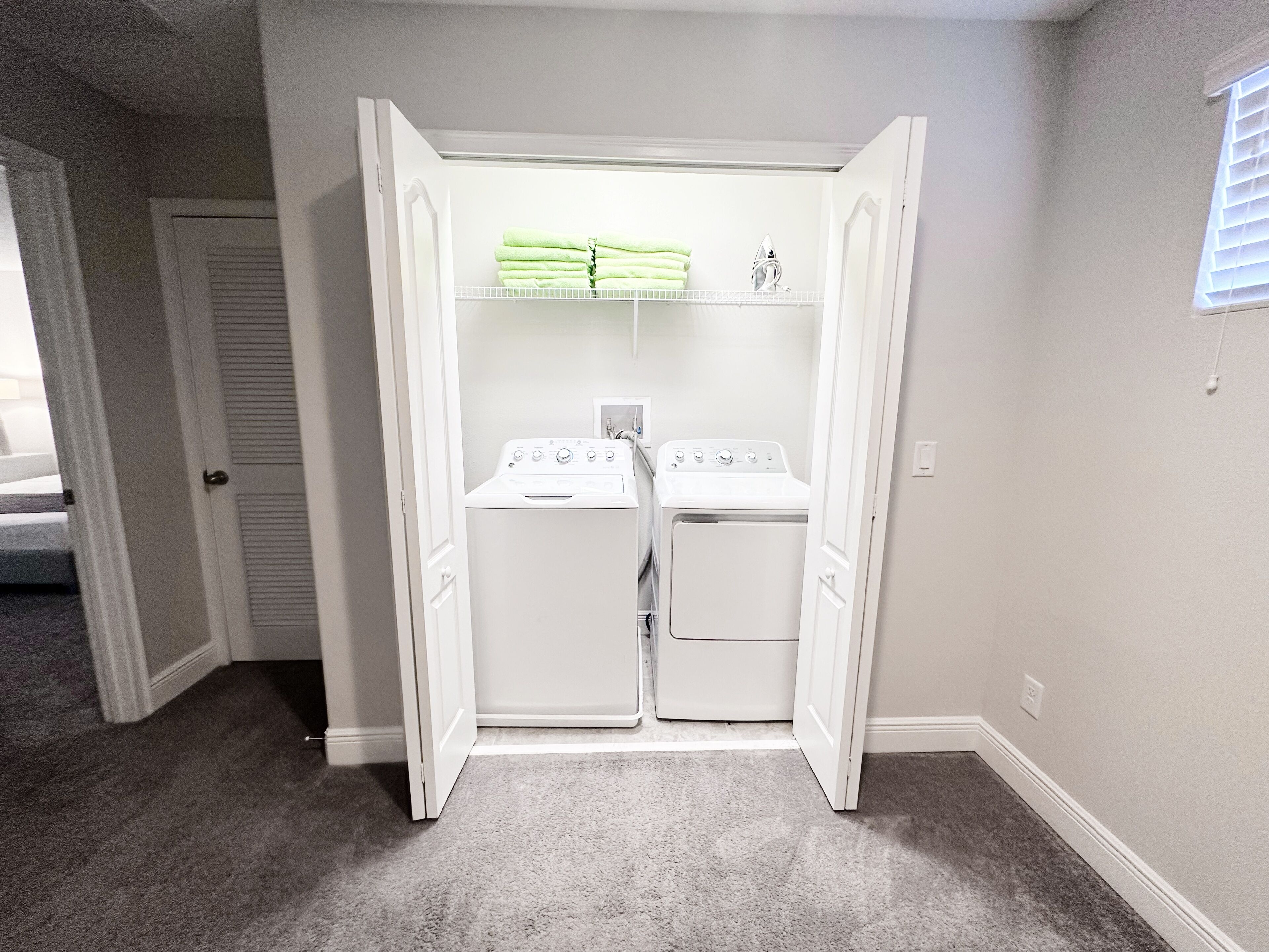 A walk-in closet with two white dryers, a hanging rack with folded towels, and a door leading to another room.