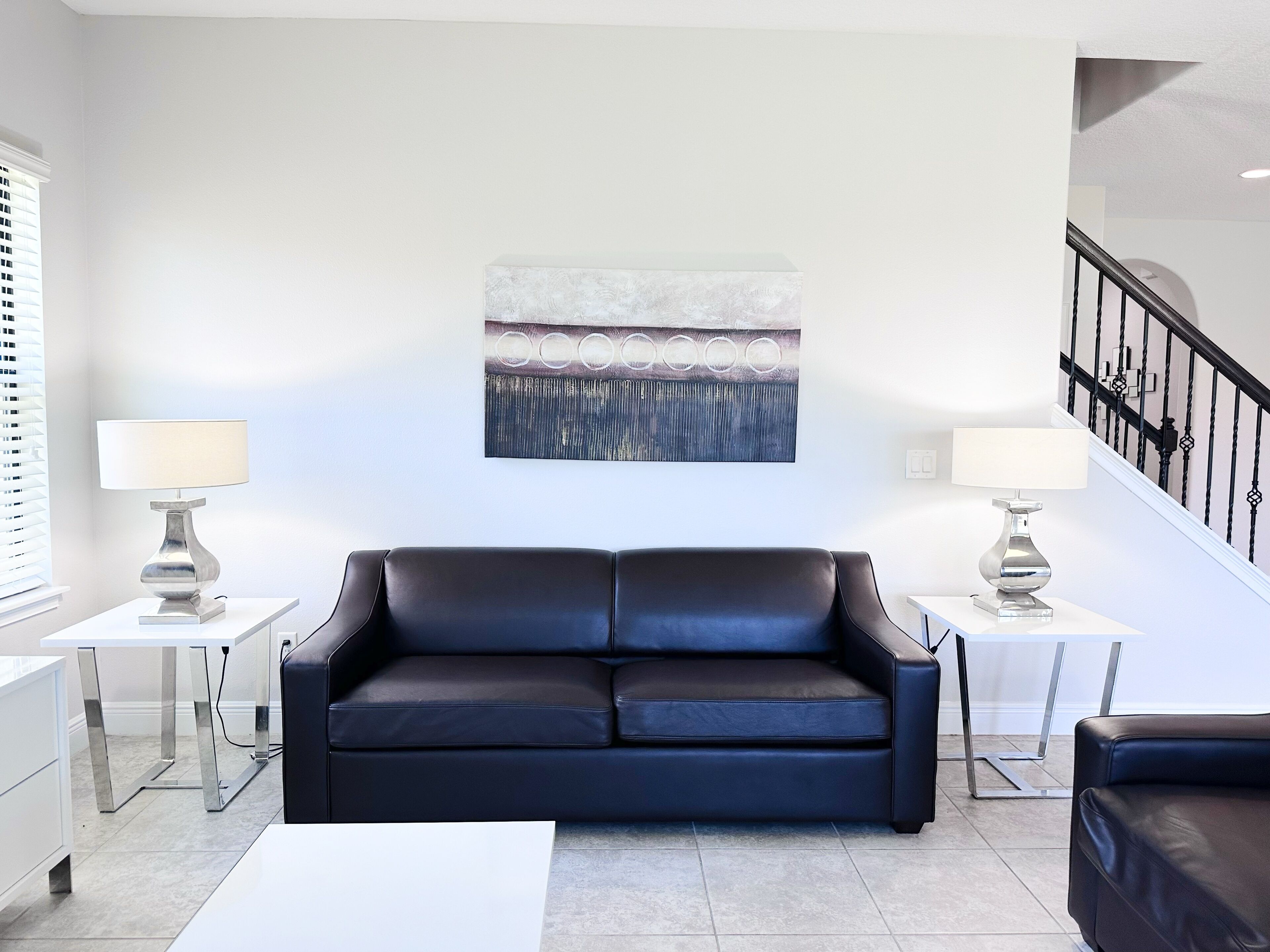 A modern living room with a black leather sofa, two side tables with lamps, a large abstract painting on the wall, and a staircase with black railings.