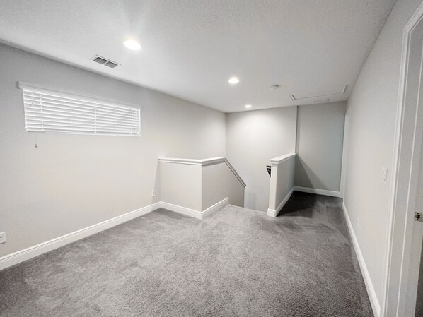 Una habitación con piso alfombrado, paredes blancas, una ventana con persianas y una puerta que conduce a otra habitación.
