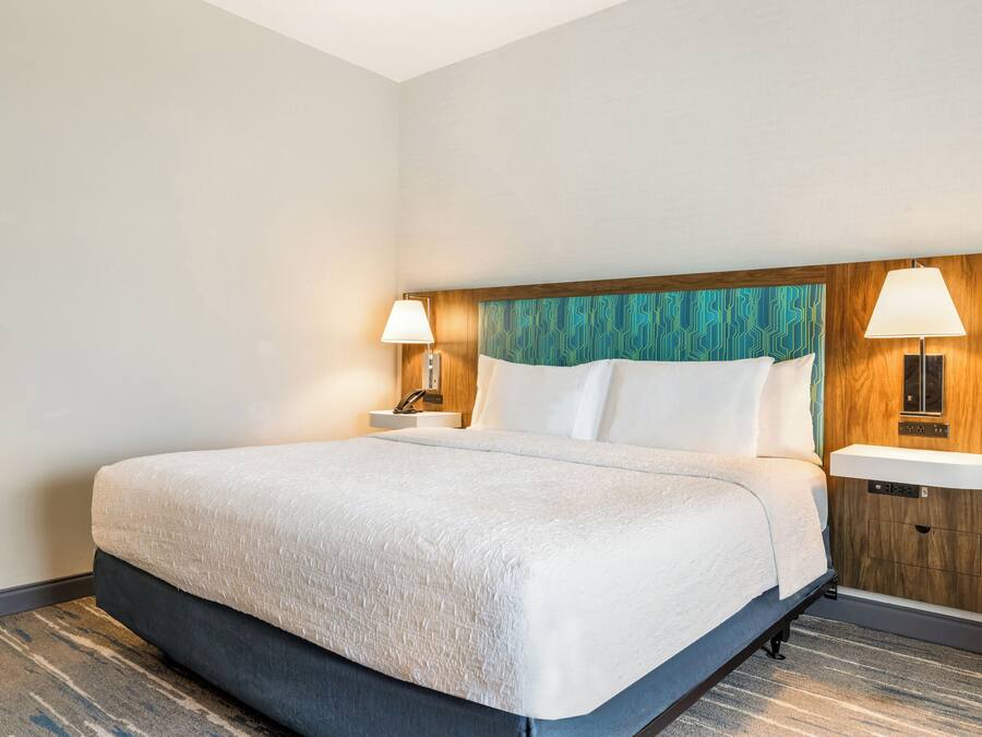 A hotel room with a large bed, two bedside tables with lamps, a wooden headboard, and a built-in shelf.