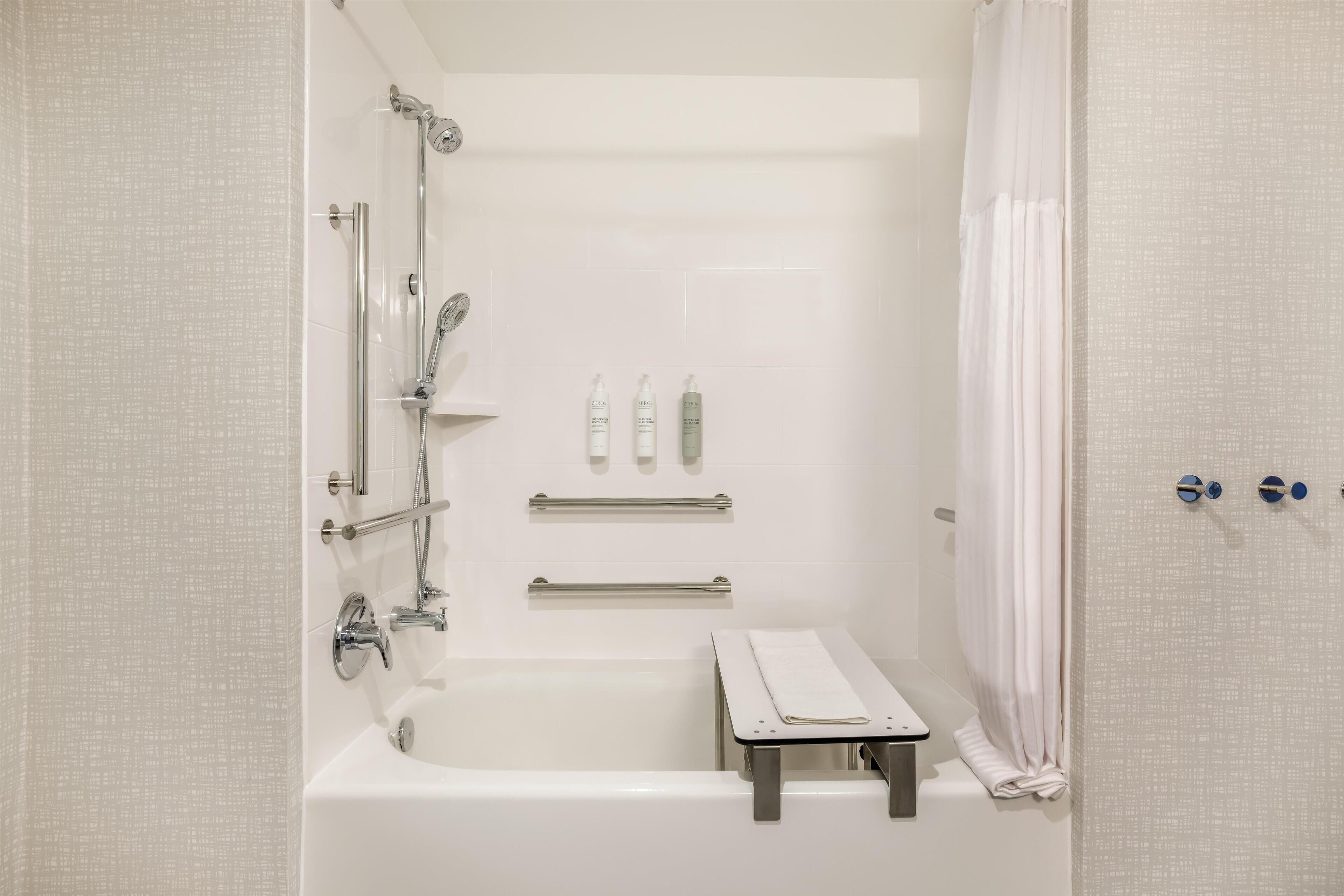 A modern bathroom with a bathtub, shower, and handrails. There are toiletries on the wall and a towel rack.