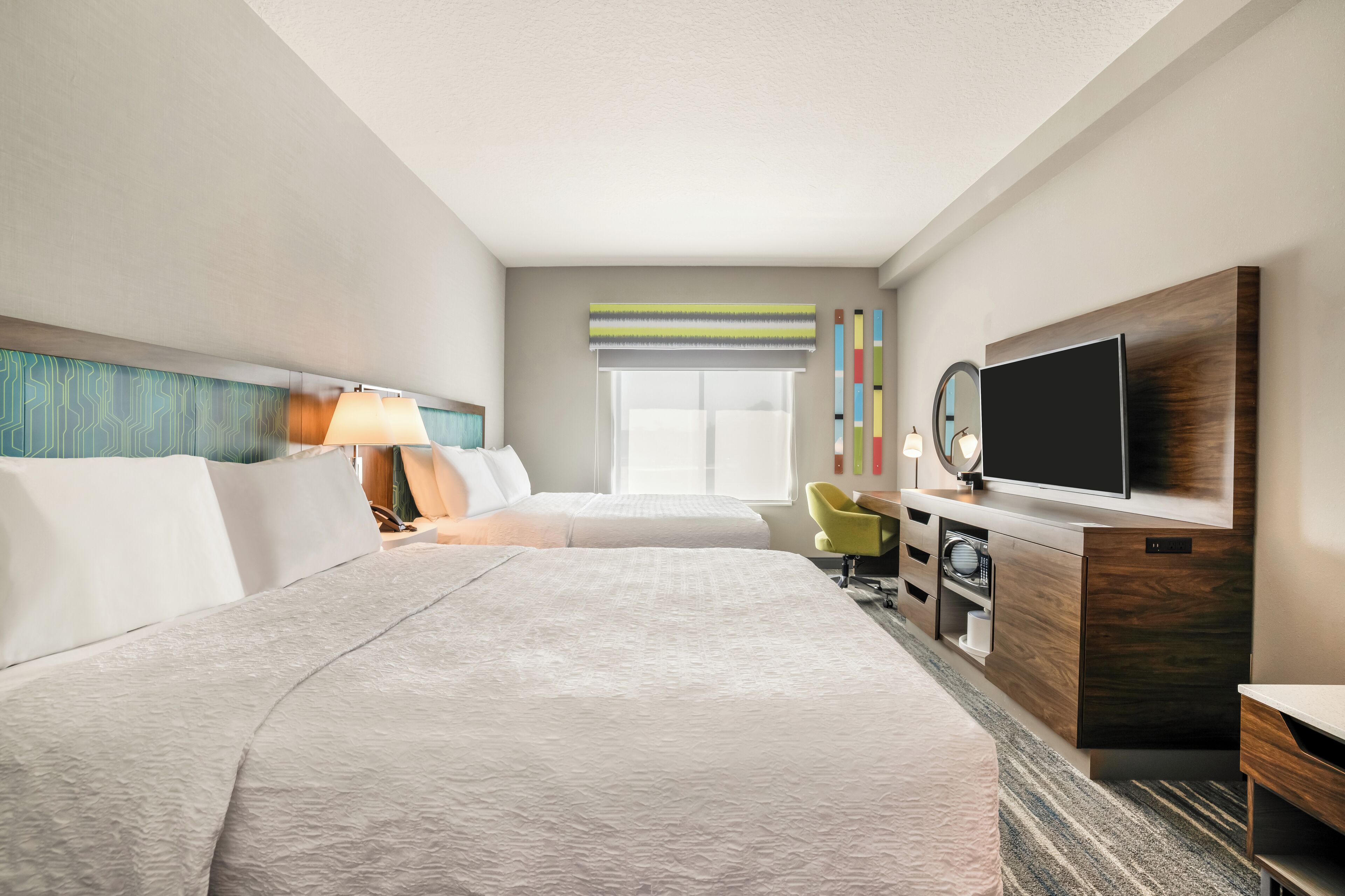 A hotel room with two beds, a TV, a desk, and a window with blinds.