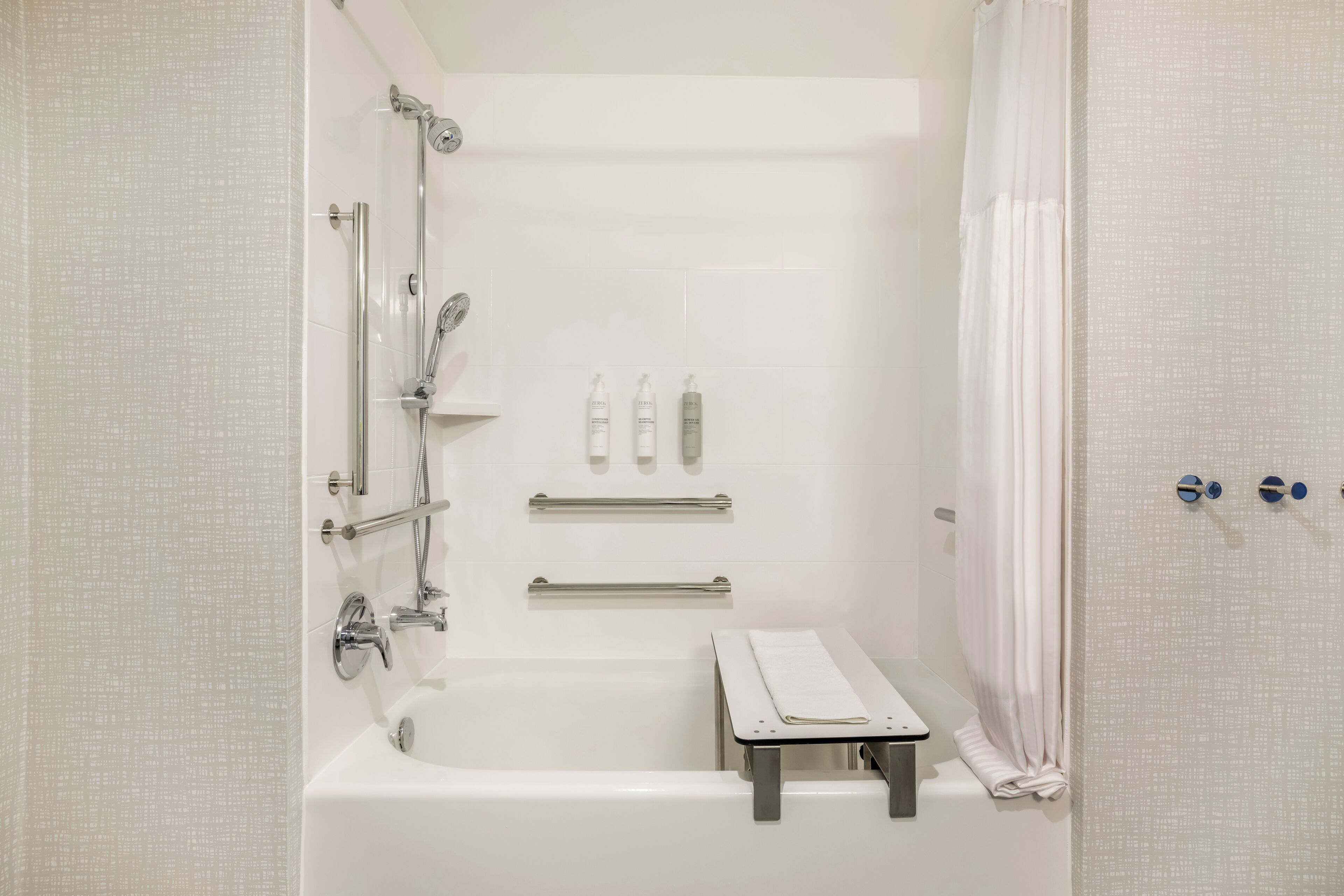 A modern bathroom with a bathtub, shower, and toilet. There are handrails, a towel rack, and toiletries on the wall.