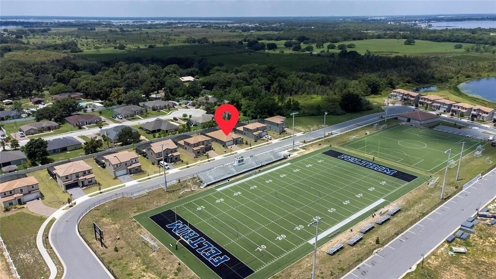 An aerial view of a football field with a stadium seating area, surrounded by residential houses and a road.