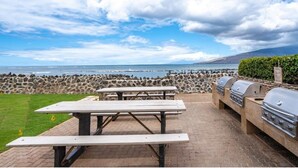 Een terras buiten met picknicktafels en een barbecue, met uitzicht op een rotsachtige kustlijn en de oceaan.