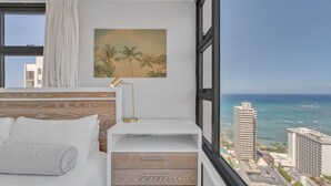 A hotel room with a bed, a desk with drawers, a lamp, and a painting of palm trees. - Beach Oasis with Ocean & City Views with FREE Parking! (Honolulu)