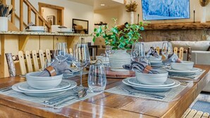 Mesa de jantar posta com pratos, copos e talheres, com um arranjo de plantas no centro e um quadro na parede.