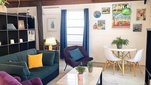 A living room with a sofa, armchairs, a coffee table, and a dining table with chairs. There is a bookshelf, a lamp, and artwork on the walls.