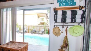 A room with a sliding glass door leading to a pool area, a wicker chest, and a sign that reads "The Captain Is In".