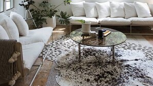 A spacious living room with a high ceiling, large windows, a round glass coffee table, and a white sofa.