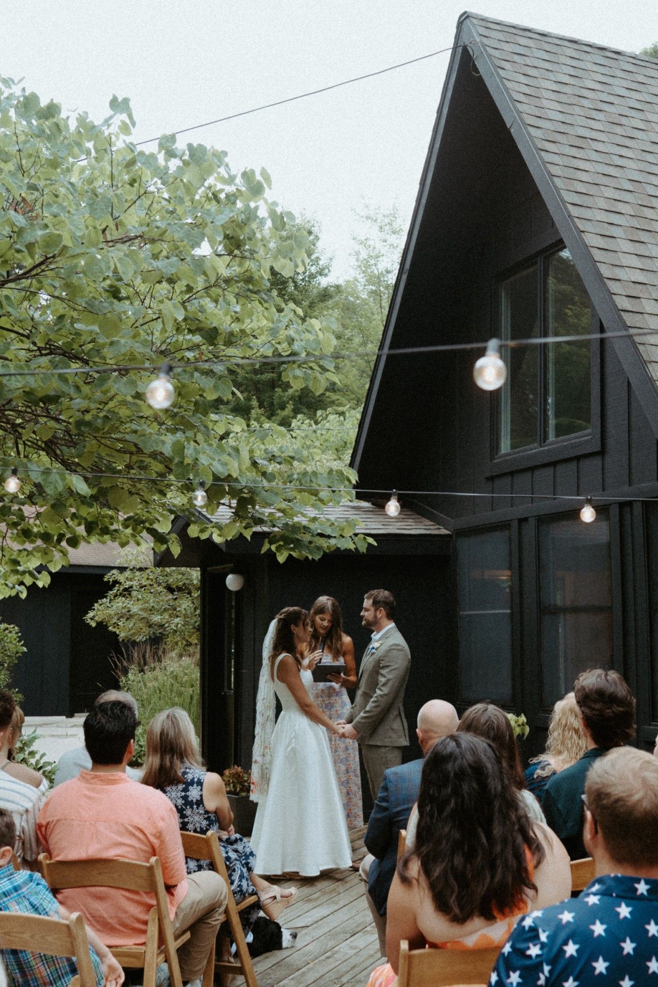 An outdoor wedding ceremony with guests seated on wooden chairs, a couple exchanging vows, and a building with a sloped roof in the background.