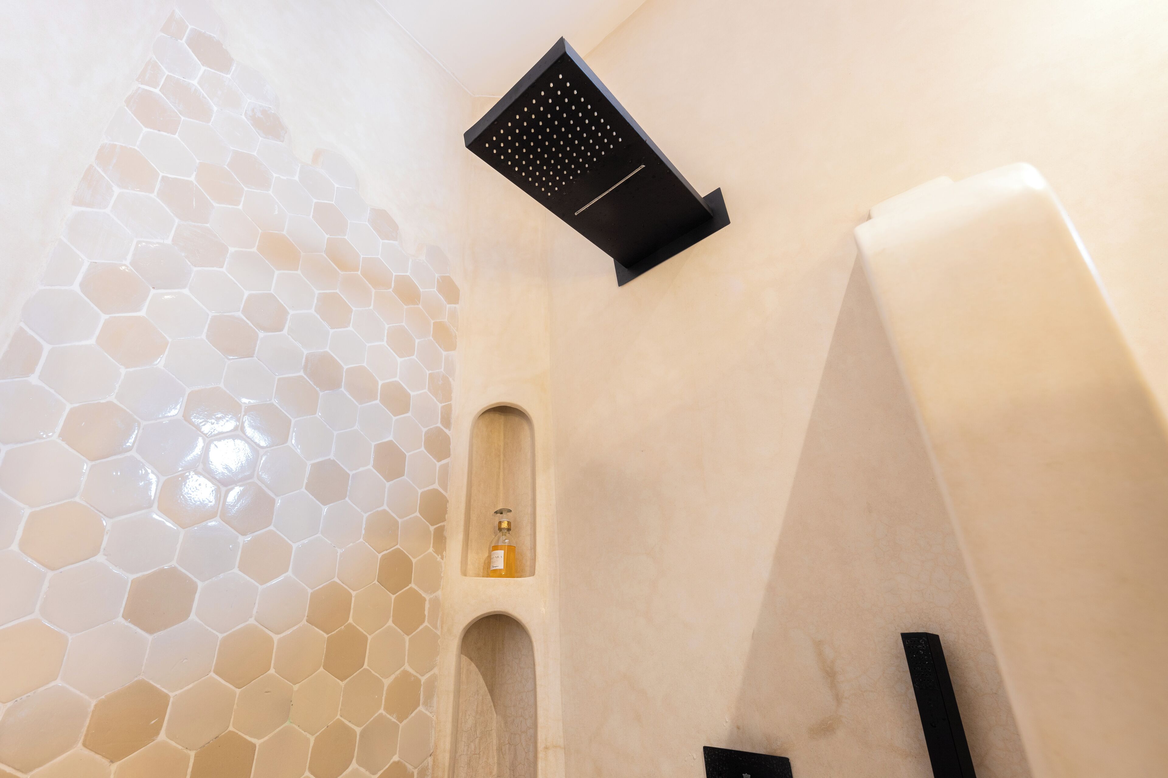 Un baño con pared de azulejos hexagonales, ducha con cabezal negro y una pequeña estantería con una botella.