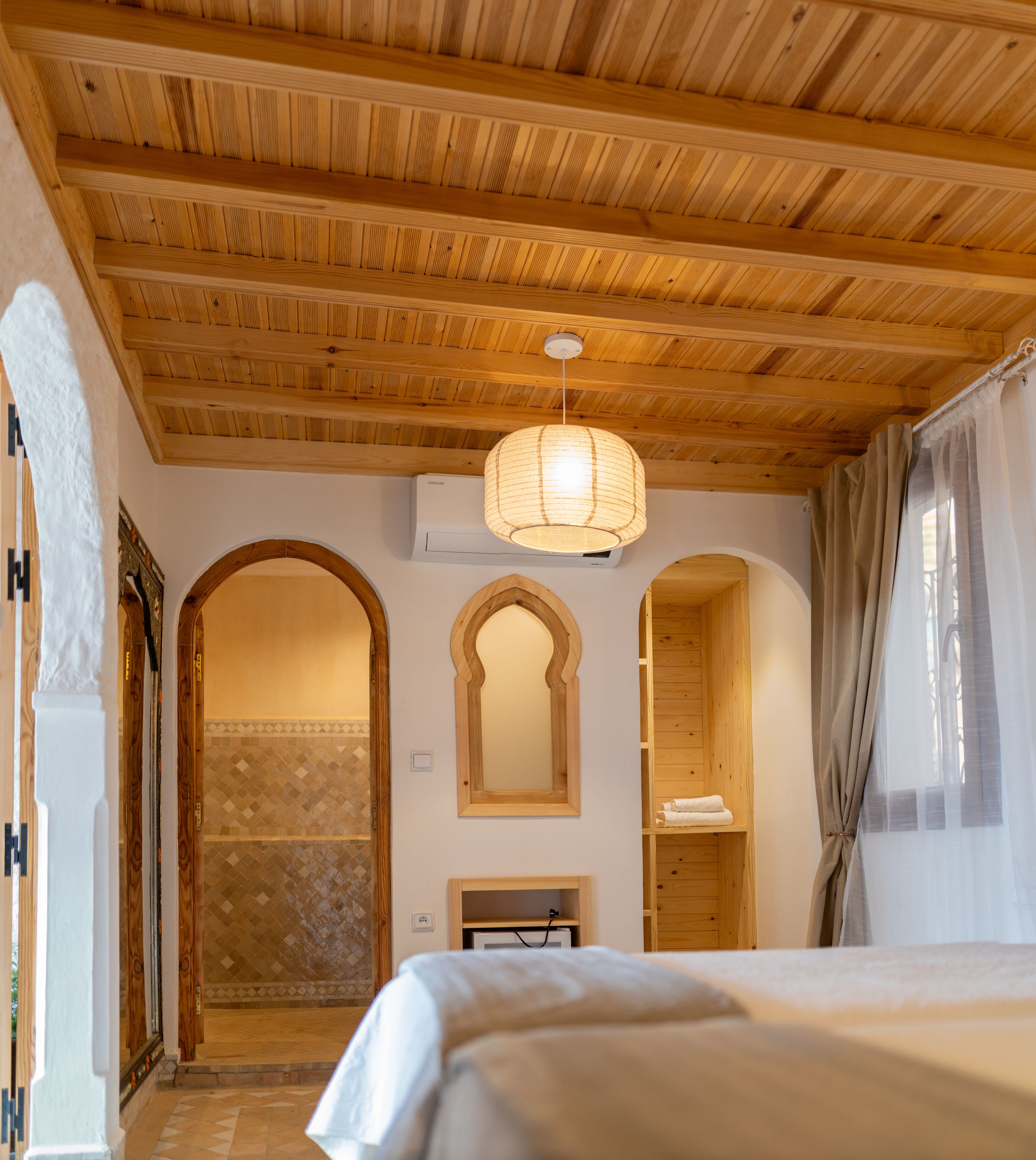 A bedroom with wooden ceiling beams, a large bed, a hanging light fixture, and a bathroom with a tiled shower area.
