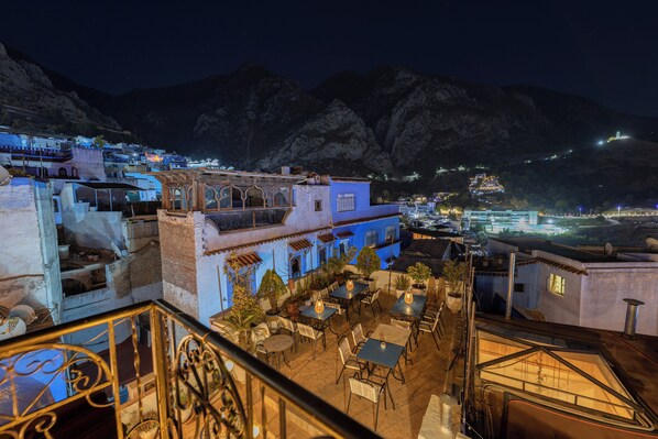 A rooftop terrace with tables and chairs, overlooking a town with illuminated buildings and mountains in the background.