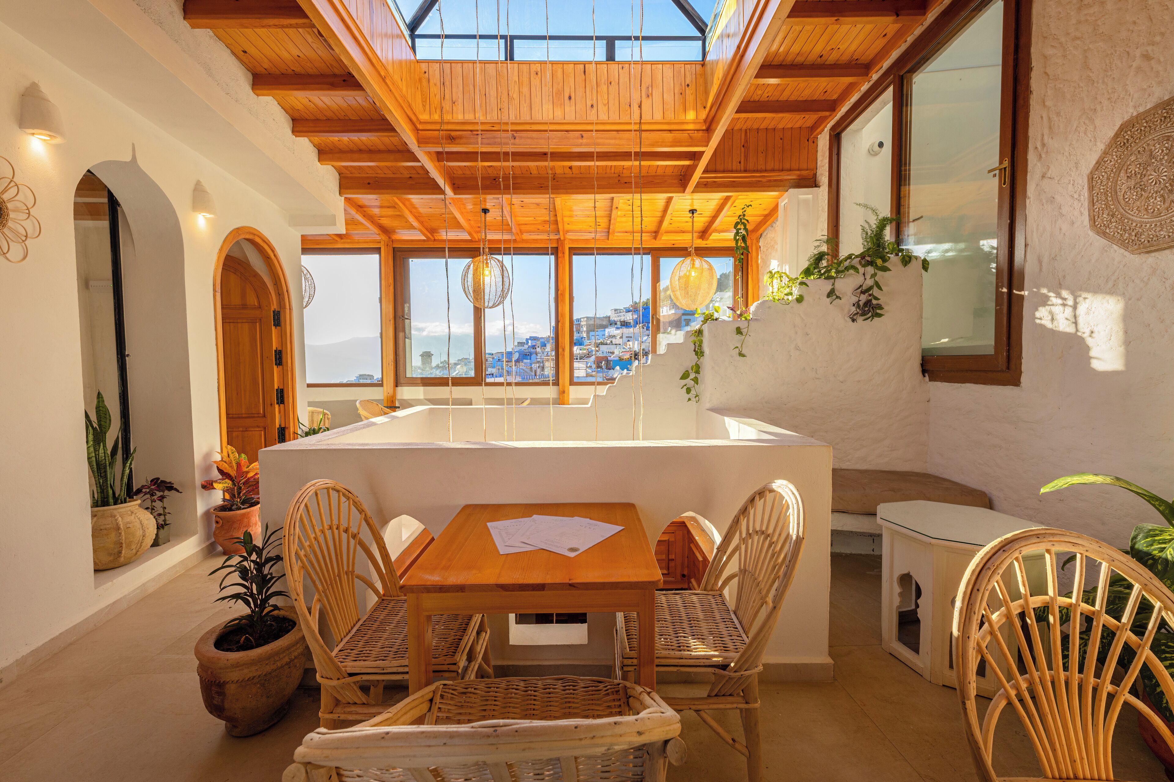 A wooden dining table with chairs, a white bench, and potted plants in a sunlit room with a view of a coastal town.